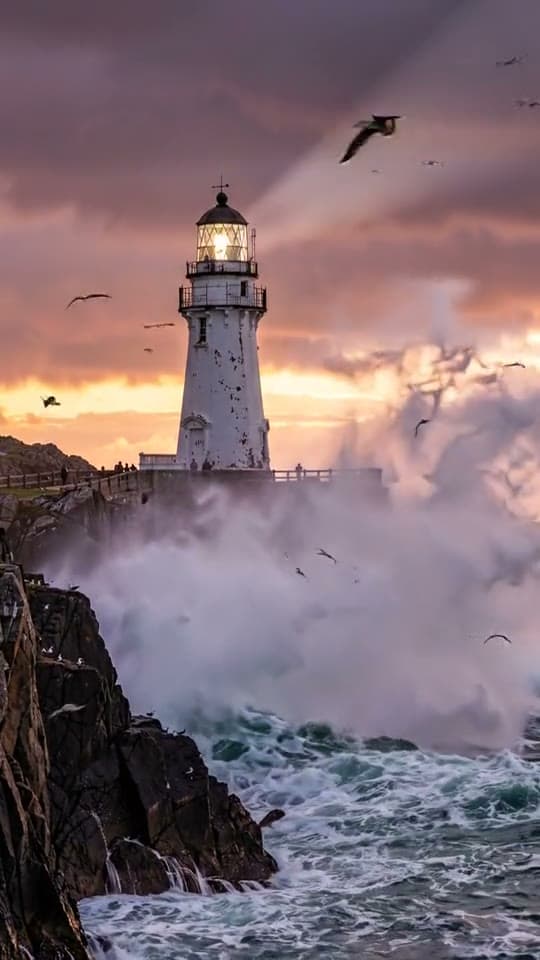 Huge waves crash against the rocks below a white lighthouse at dusk as gulls circle and the beam sweeps the sky