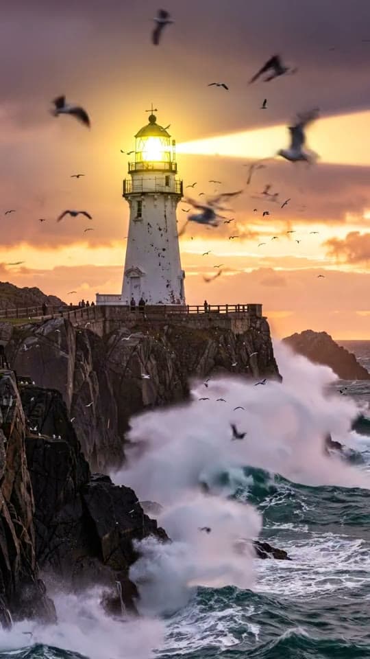 Waves crash below a white lighthouse at sunset as the beam sweeps the sky, ending on the frame it started on