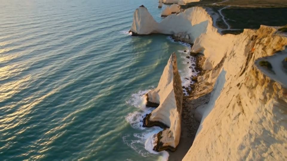A drone rises over turquoise sea and white cliffs at sunrise and tilts up to the coastline
