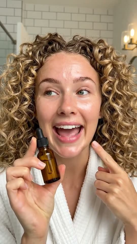 A woman with curly hair in her bathroom holds a serum bottle up to the phone and talks about it